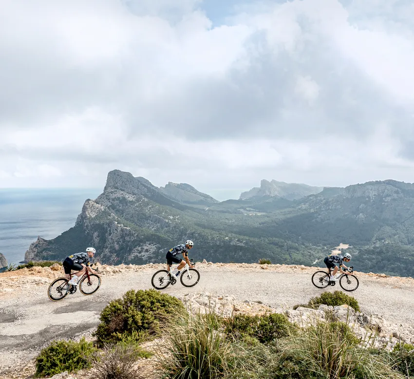 Tre ciclisti in abbigliamento sportivo pedalano su una strada lungo un paesaggio costiero con montagne e mare sullo sfondo.